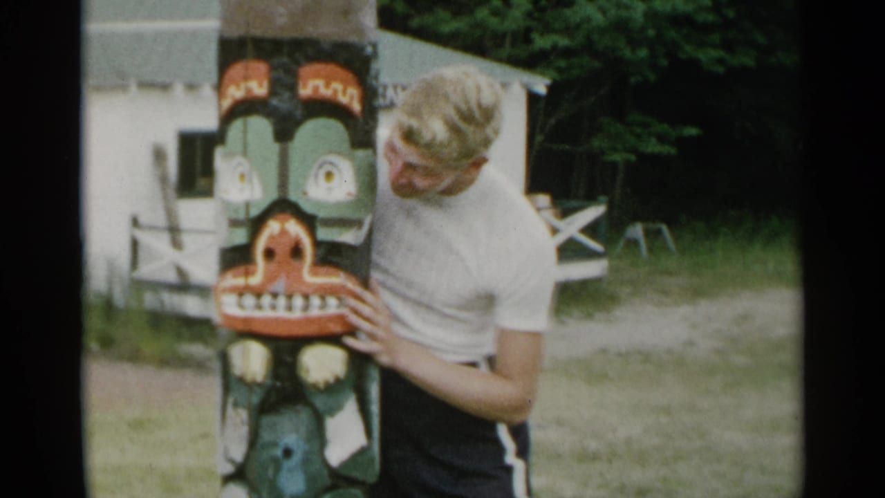 Young man posing beside a painted carved post near a rustic building.