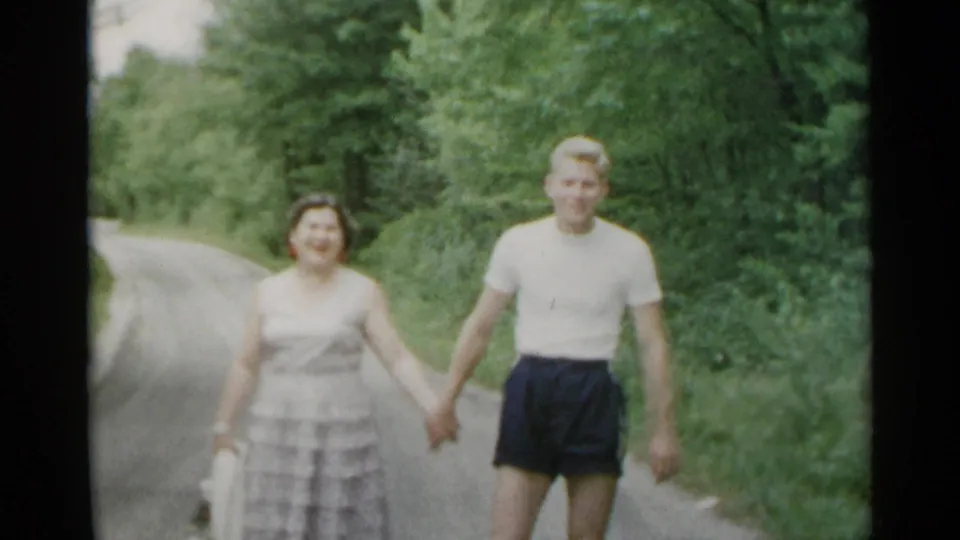 A woman in a light dress and a young man in shorts walk hand-in-hand on a wooded road in metadata-supported 1954 New York.