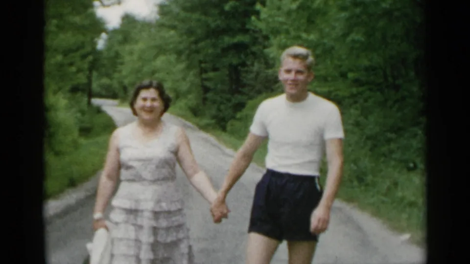 A smiling woman and young man continue toward the camera on a curving wooded road in 1954 New York home-movie footage.
