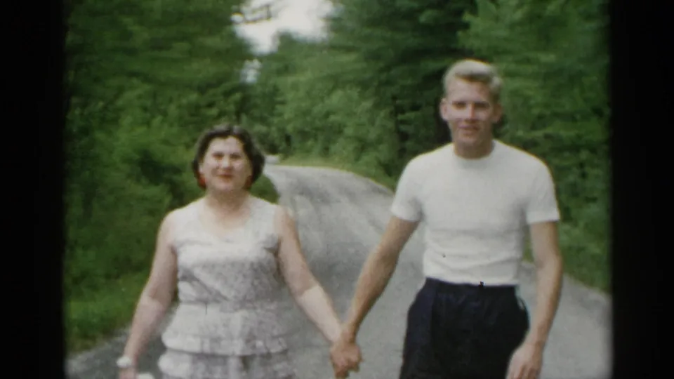 The woman and young man are framed closer as they hold hands on a wooded road, with soft 8mm color and visible faces.