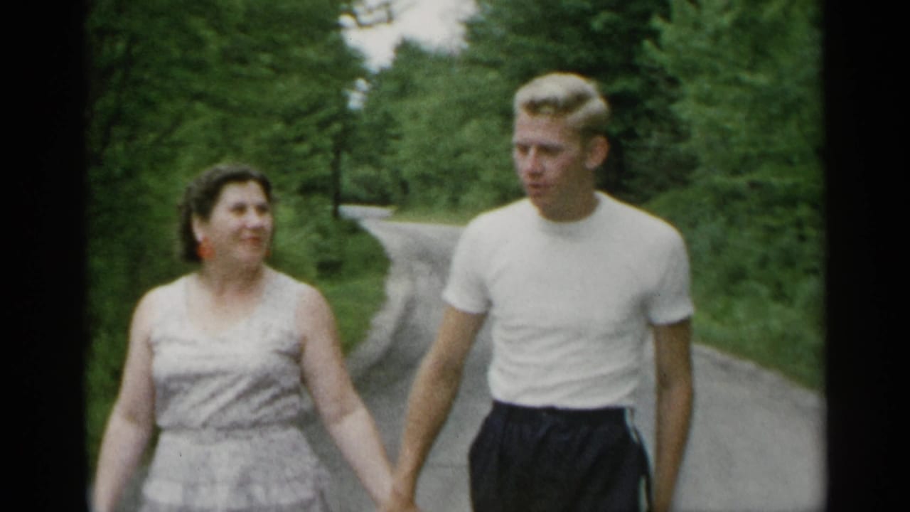 In 1954 New York home-movie footage, a woman in a sleeveless dress and a blond man in a white t-shirt walk hand-in-hand along a tree-lined path.