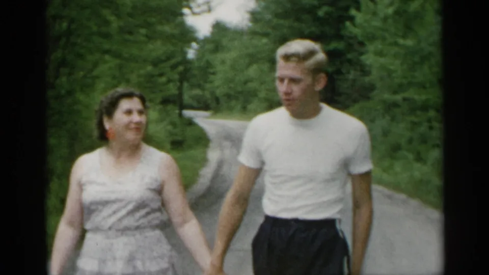 In 1954 New York home-movie footage, a woman in a sleeveless dress and a blond man in a white t-shirt walk hand-in-hand along a tree-lined path.
