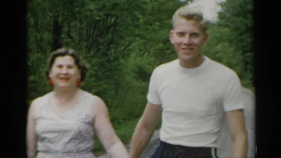 The couple approaches the camera in a wooded New York park, with the woman smiling and the man in a white t-shirt beside her.