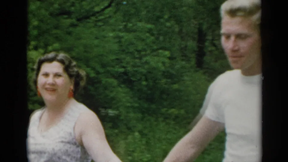 A closer 1954 home-movie view shows the woman smiling near the camera while the man walks partly cropped at her side.