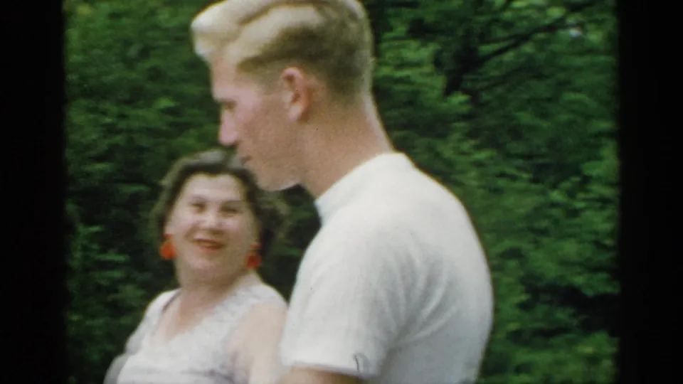 The man is seen in profile while the woman looks toward him, both walking through a leafy park in metadata-supported 1954 New York footage.