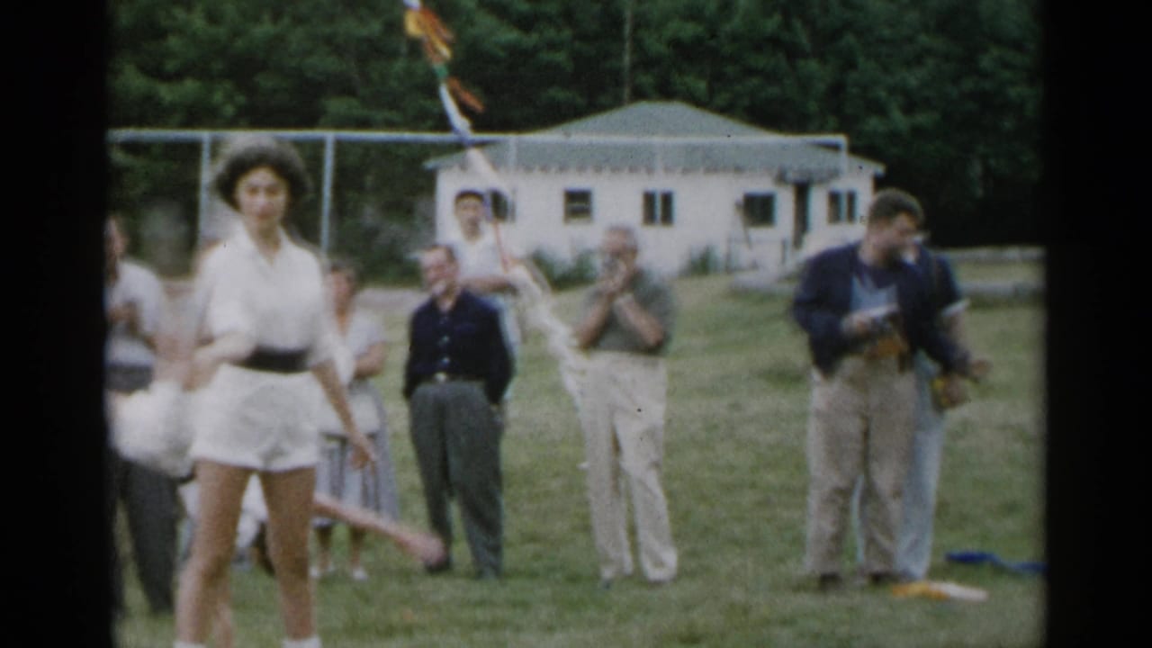 Teen in white tennis outfit stands on a grassy field while adults watch behind her.