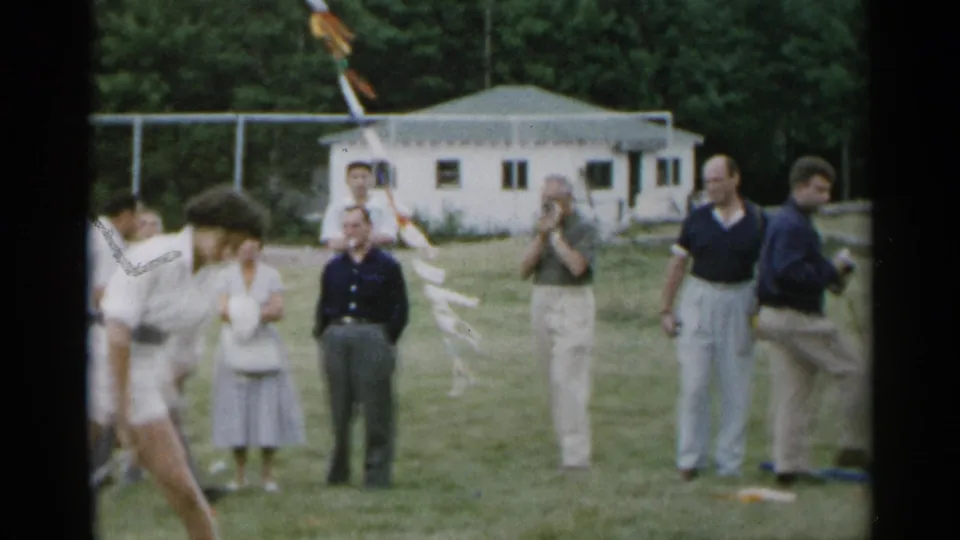 1954 New York home-movie frame with spectators watching from a grassy field while the tennis-clad teen passes partly out of view.