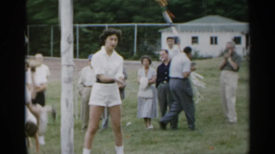 1954 New York home-movie frame showing a teen in tennis attire near a pole with adults standing and clapping in the background.