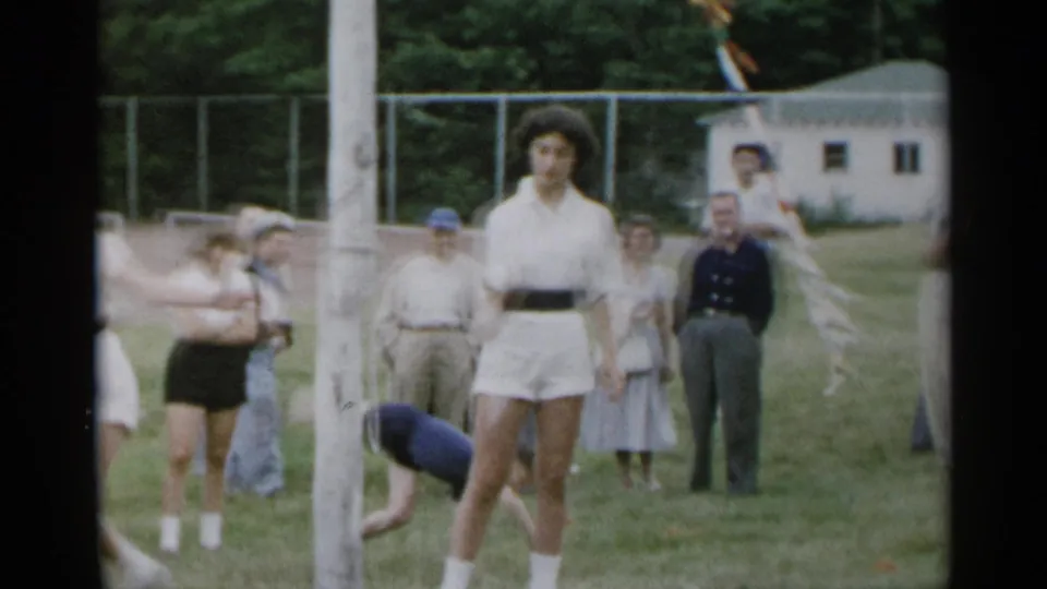 1954 New York home-movie frame with the teen centered in a white tennis outfit, spectators behind her, and a person bending near the grass.
