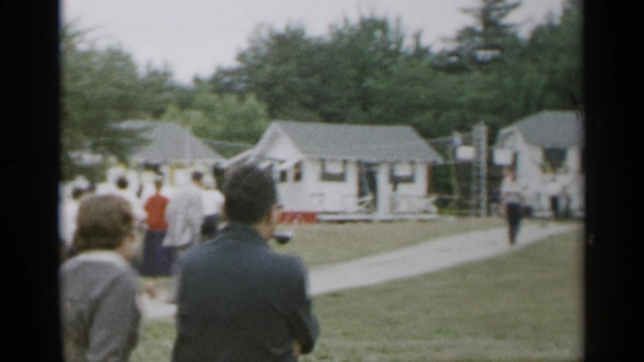 Adults stand on a lawn facing small white cottages and a sidewalk.