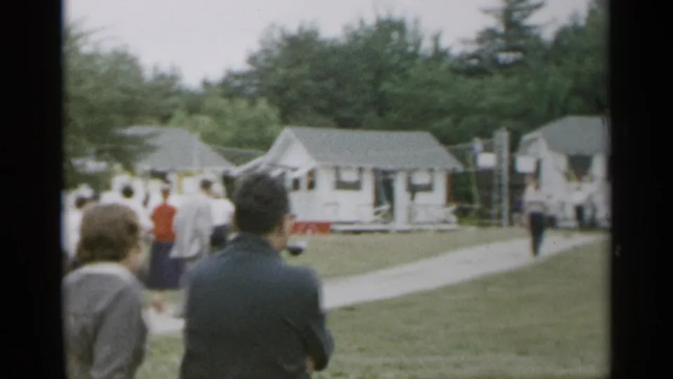 1954 New York home-movie frame showing adults on a grassy lawn looking toward small white cottages with trees behind them.