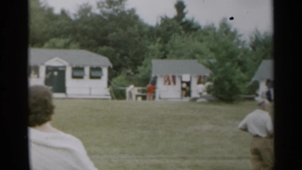 1954 New York home-movie frame showing a grassy cottage area with scattered adults and trees in the background.