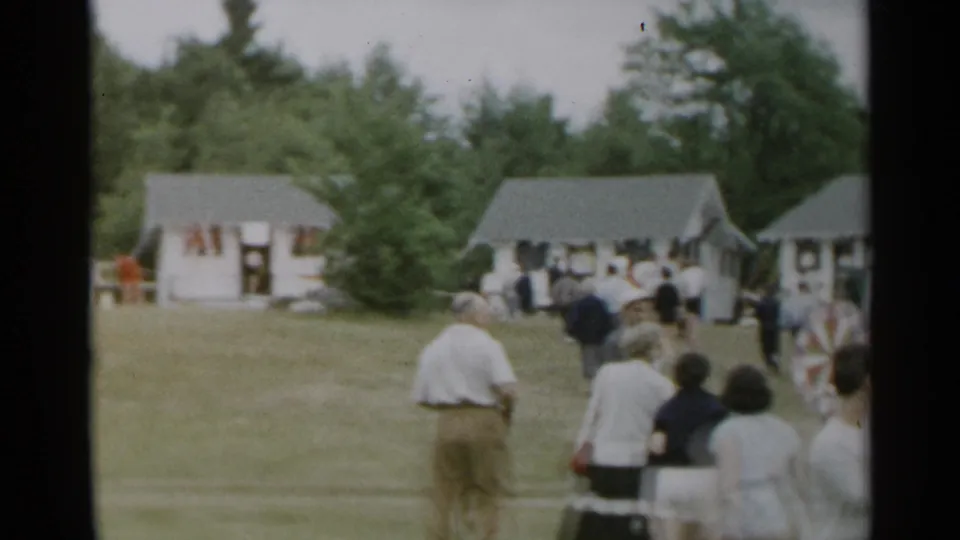 1954 New York home-movie frame showing adults crossing a grassy lawn toward small white cottages beneath a tree line.