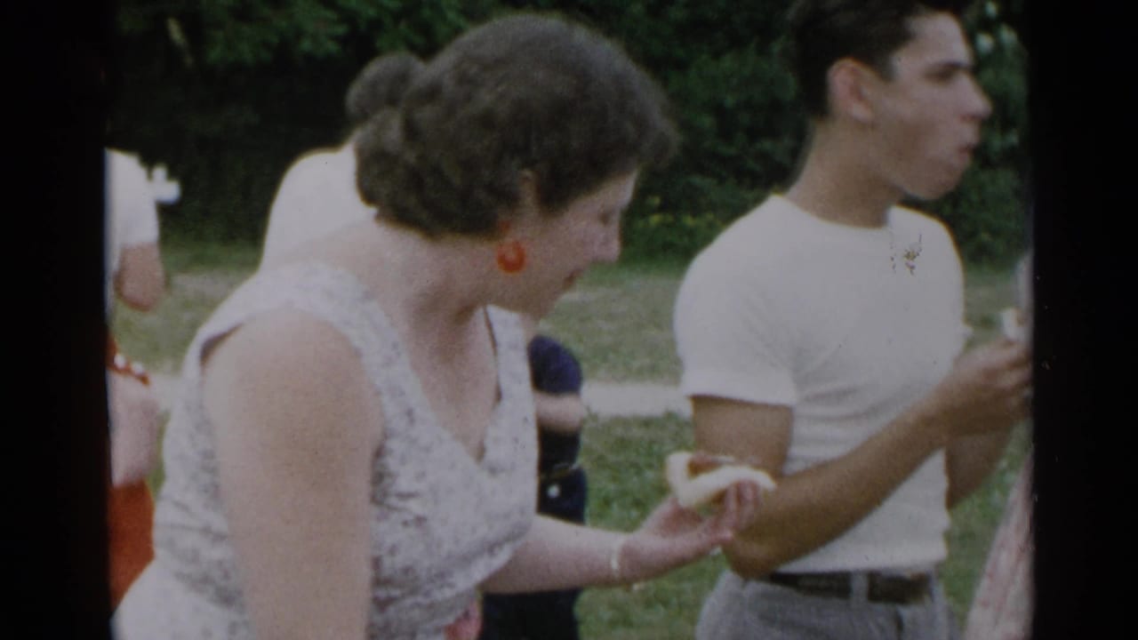 Woman holding a hot dog beside a man in a white T-shirt at an outdoor cookout.