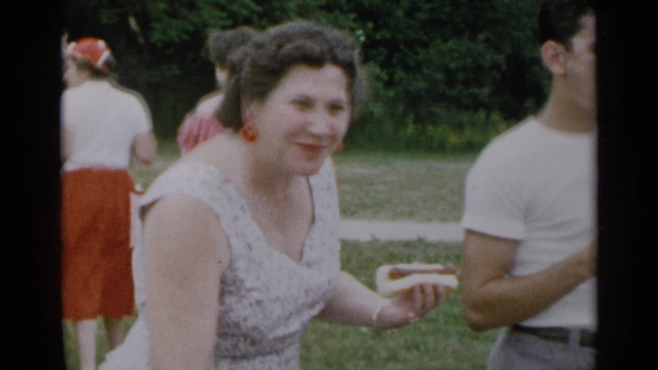 Woman smiles while holding a hot dog near other adults at a grassy outdoor gathering.