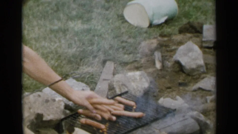 1954 New York home-movie frame showing hot dogs on a grill over coals, with smoke, stones, grass, and a hand reaching near the fire.