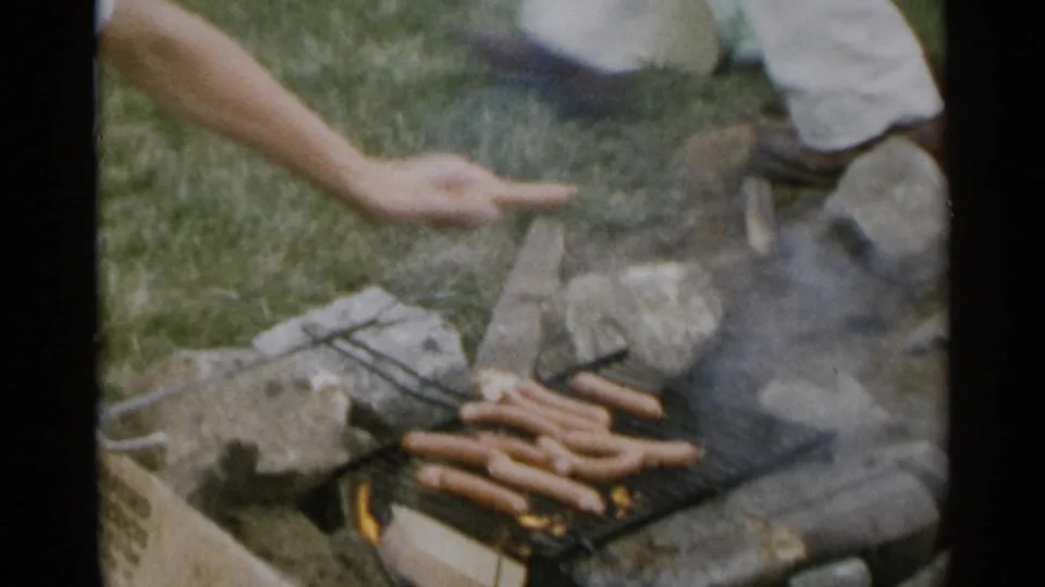 1954 New York home-movie frame showing hot dogs grilling over a small campfire with smoke, stones, grass, and an adult hand nearby.
