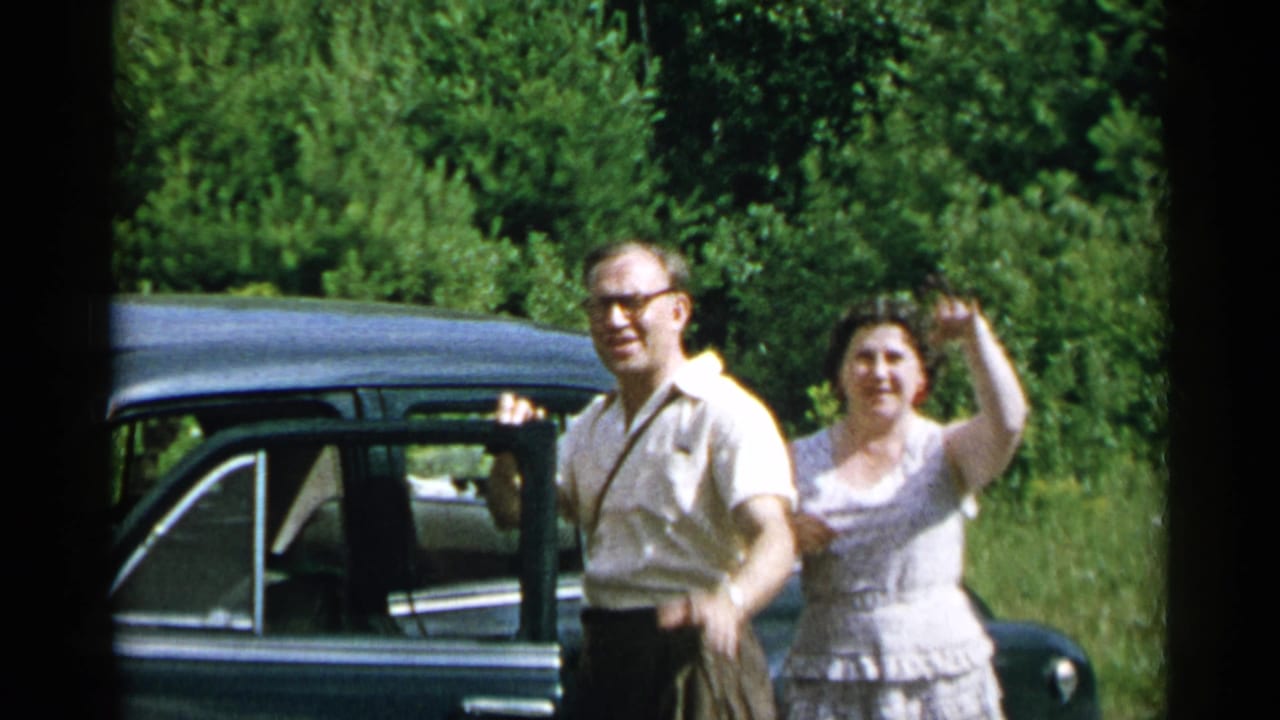 Adults stand beside a dark sedan on a sunlit rural road, with one woman waving.