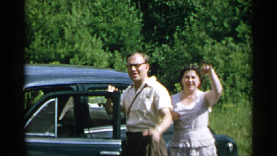 1954 New York home-movie frame showing adults beside a dark sedan on a tree-lined rural road, with a woman raising her hand.