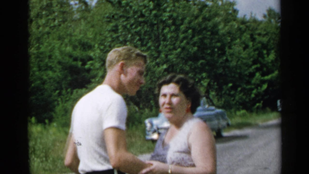 1954 New York home-movie frame of a man and woman standing close on a rural roadside, with older cars and dense trees behind them.