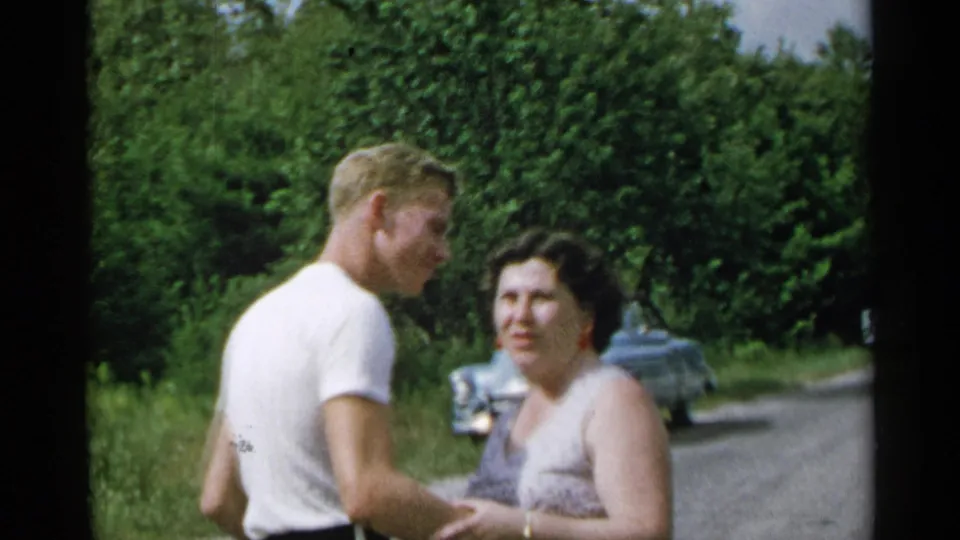 1954 New York home-movie frame of a man and woman standing close on a rural roadside, with older cars and dense trees behind them.