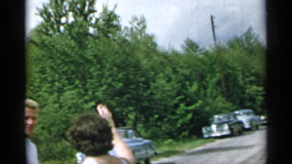 1954 New York home-movie frame with a woman seen from behind waving toward the road, where older cars are parked near dense greenery.