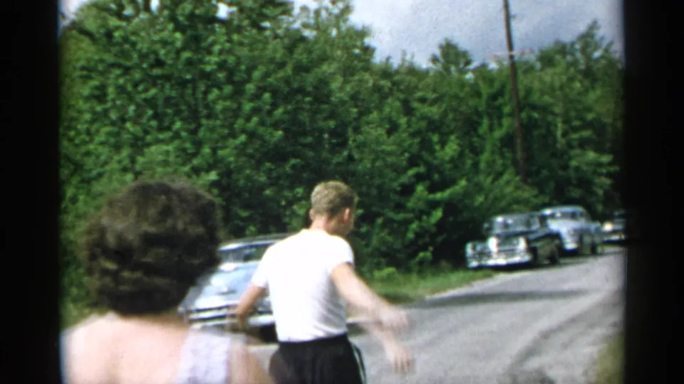 1954 New York home-movie frame showing a man walking away along a rural road, with a woman in the foreground and older cars parked ahead.