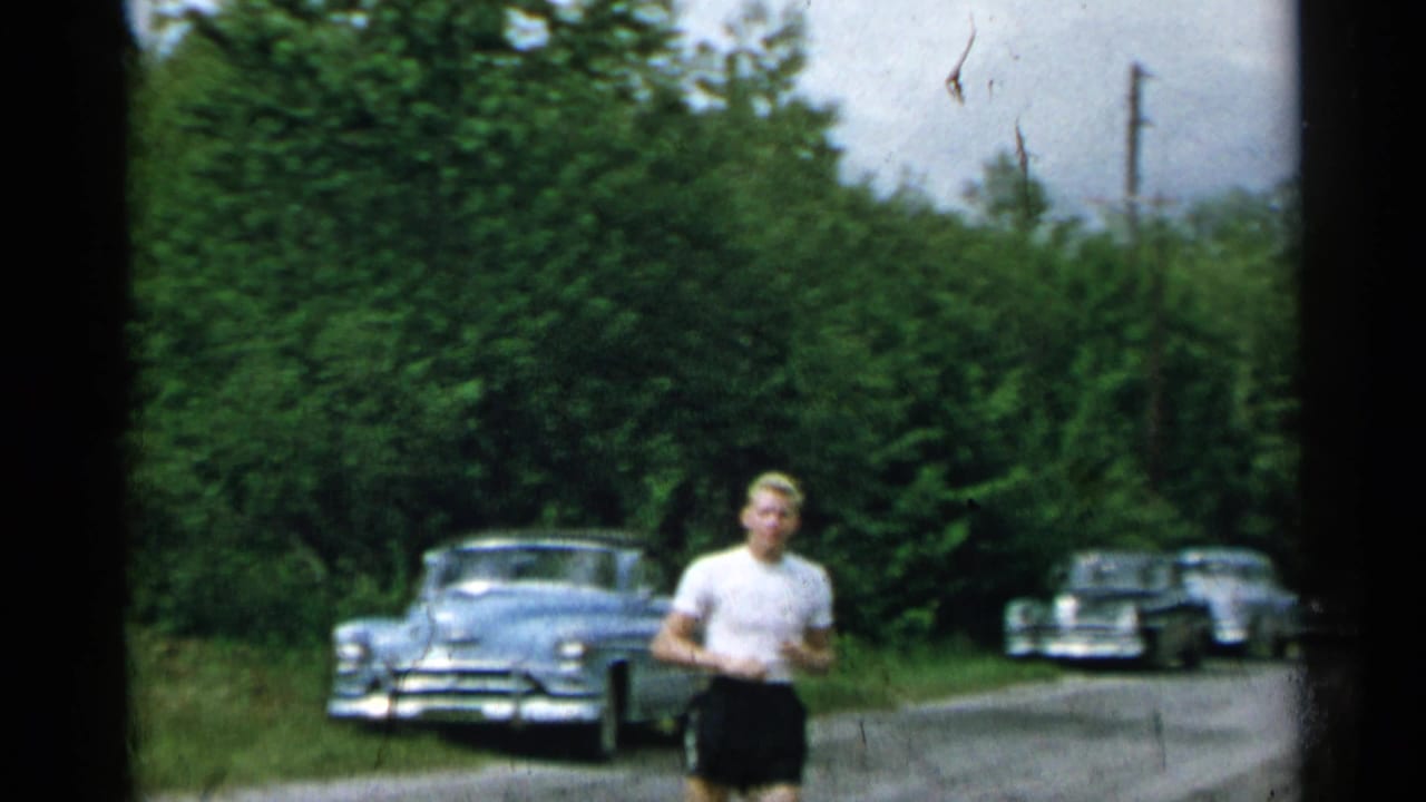1954 New York home-movie frame of a man in a white T-shirt and dark shorts walking along a quiet road with mid-century cars nearby.