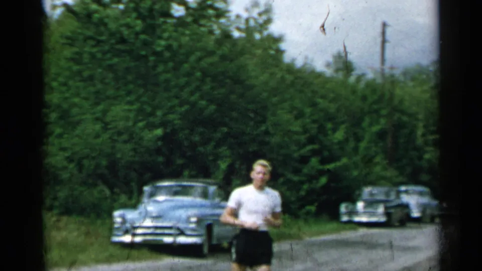 1954 New York home-movie frame of a man in a white T-shirt and dark shorts walking along a quiet road with mid-century cars nearby.