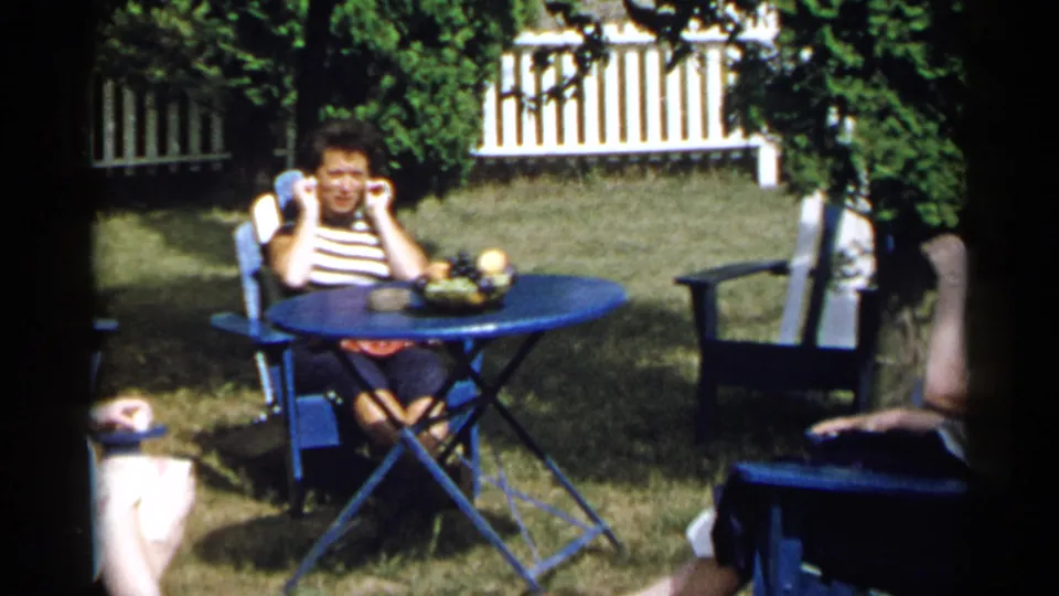 1954 New York garden scene with an adult at a blue folding table and another seated partly in frame near shrubs and a white fence.