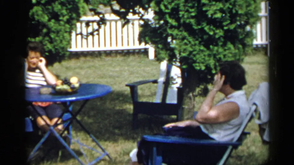 1954 New York home-movie frame of two adults seated in a garden, one beside a blue table and one reclining in a blue chair.
