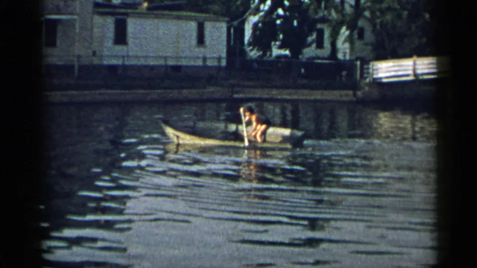 1954 New York 8mm frame of a person in dark shorts rowing a yellow rowboat past shoreline houses and trees.