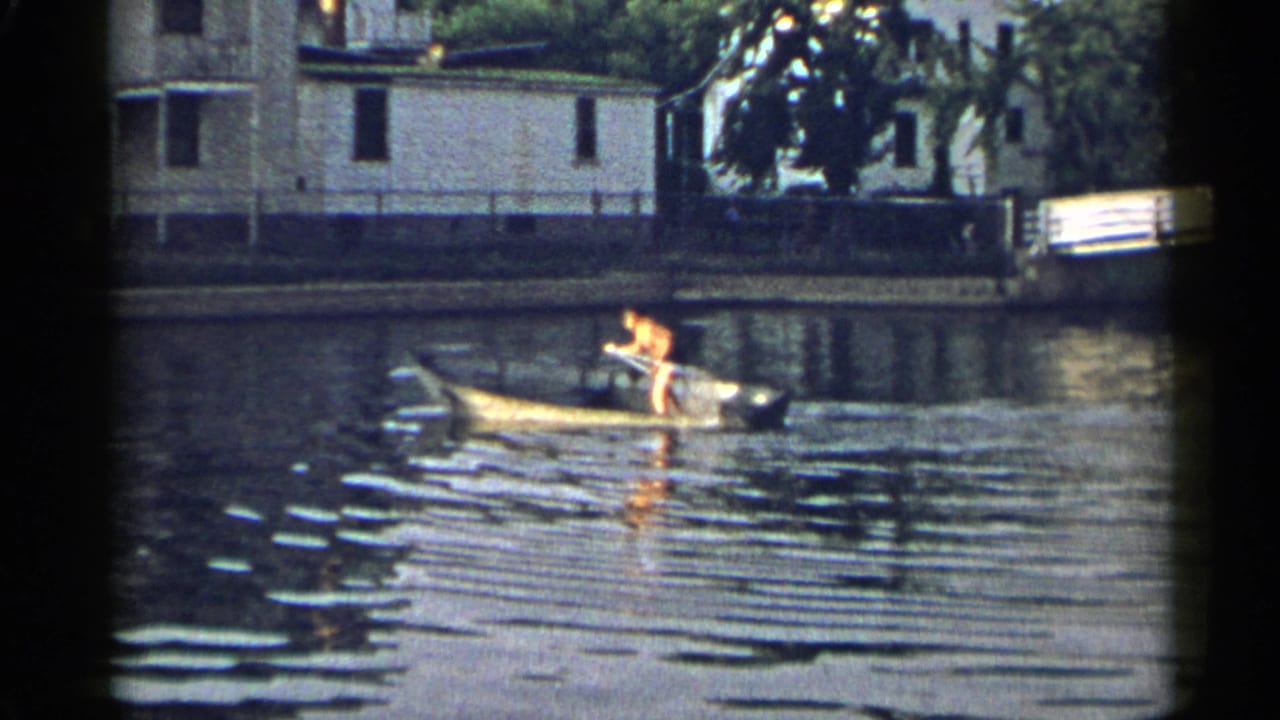 A person rows a yellow rowboat along a quiet New York waterway, with residential shoreline buildings behind.