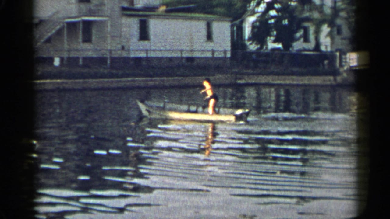 The rower appears upright in the yellow boat on calm water, with a light-colored shoreline house behind.