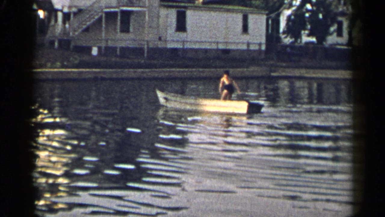 A yellow rowboat sits on a quiet waterway as a person remains aboard near residential shoreline buildings.
