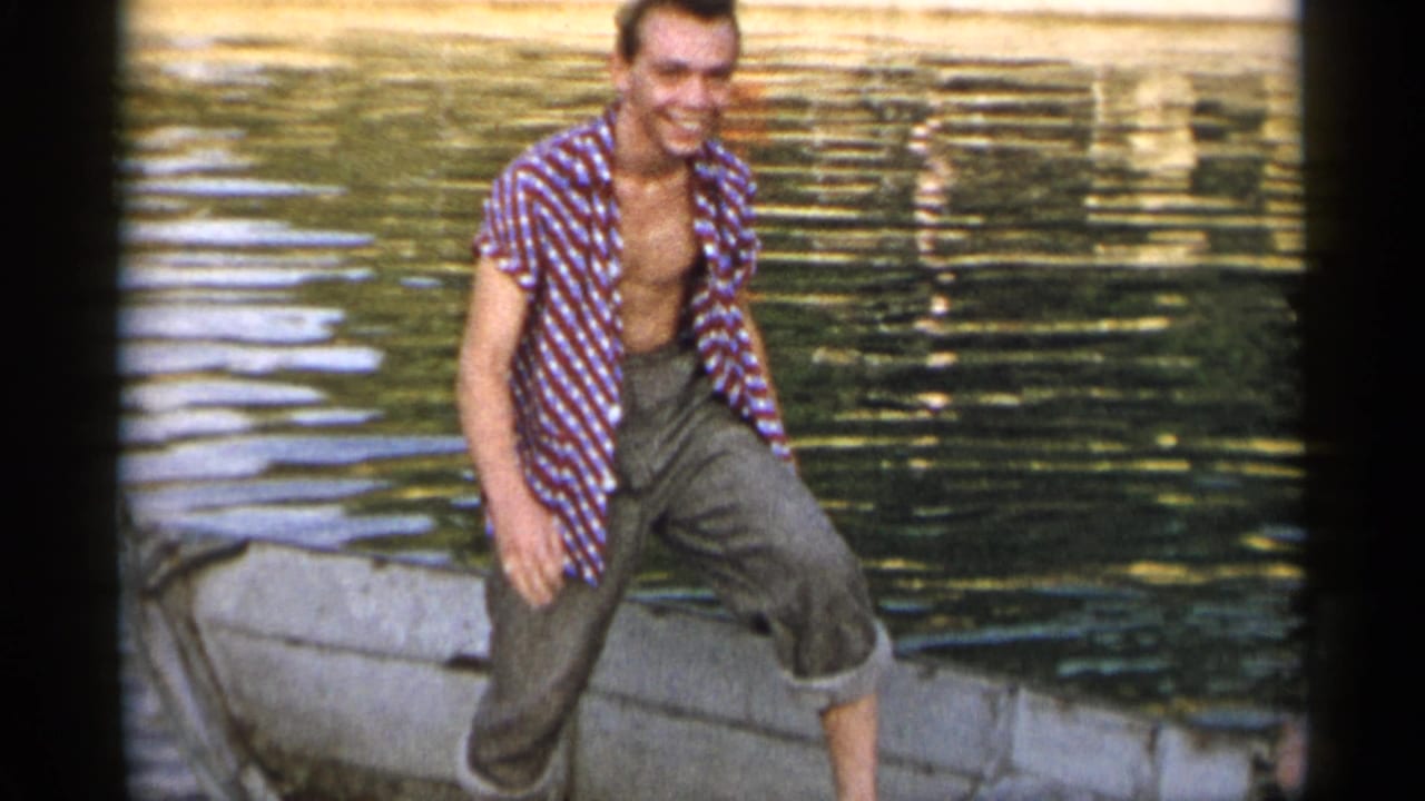1954 New York frame of a smiling adult man standing in a small boat, with rippled lake water behind him.