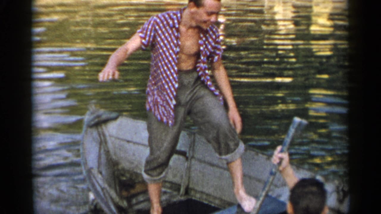 Adult man in rolled trousers balances in a small boat as another person with an oar or paddle appears at the edge of the frame.