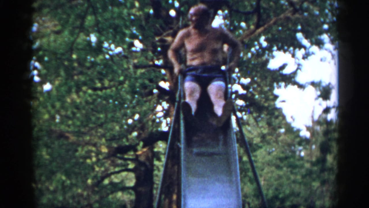Shirtless older man seated at the top of a metal slide beneath leafy trees.