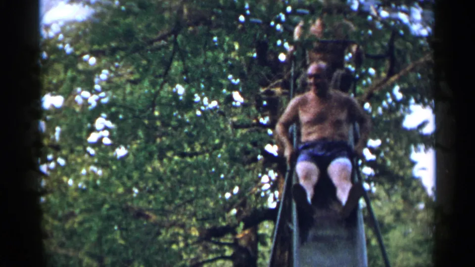 A shirtless older man rides down a metal slide while another person is partially visible above, framed by trees in 1954 New York footage.