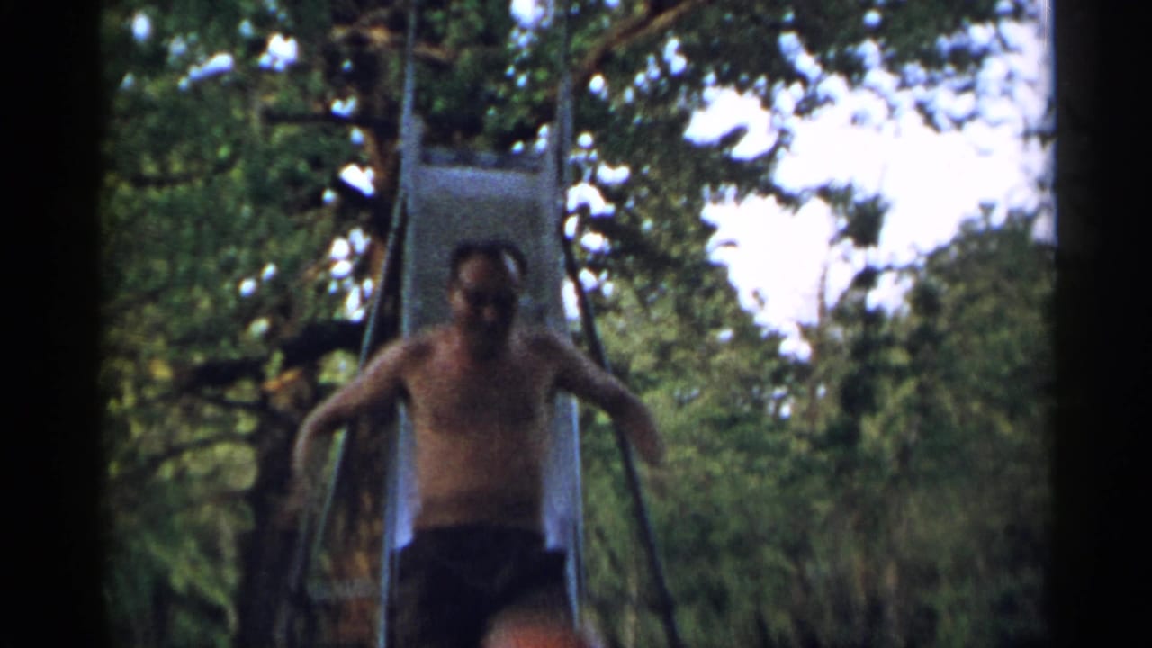 The man descends the metal slide, arms out near the rails, in soft 8mm footage with dense greenery behind him.