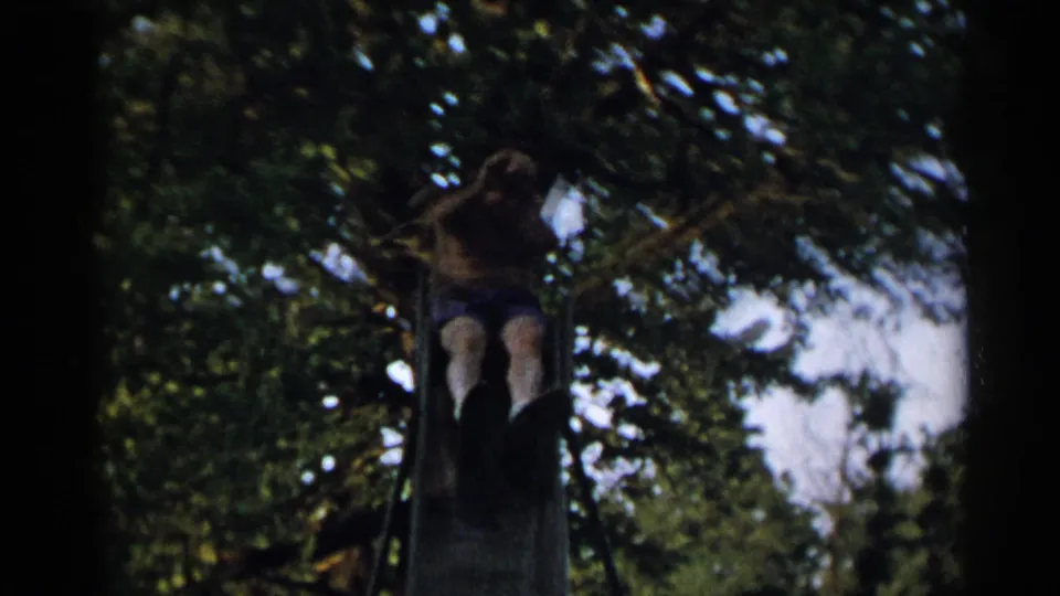 In 1954 New York home-movie footage, a shirtless older man sits near the top of a metal park slide beneath leafy trees.