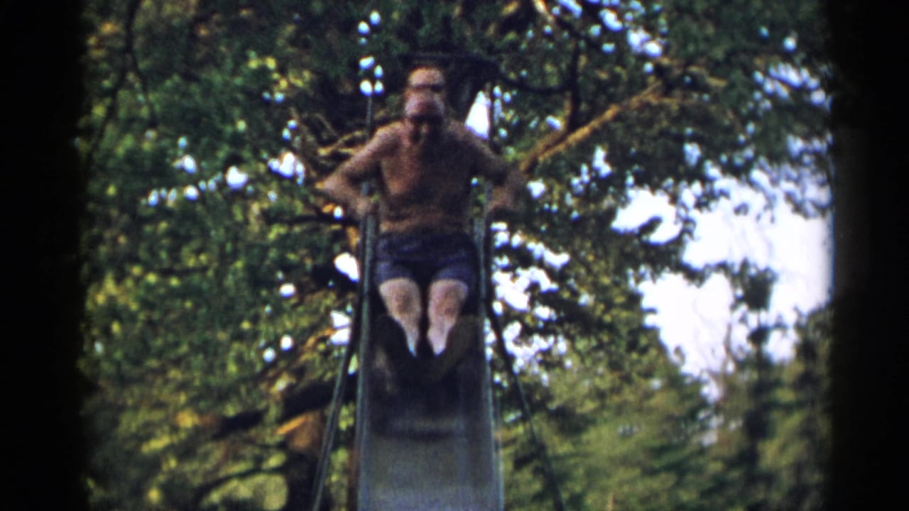 The man faces the camera from the top of the metal slide, framed by trees in the 1954 New York park footage.