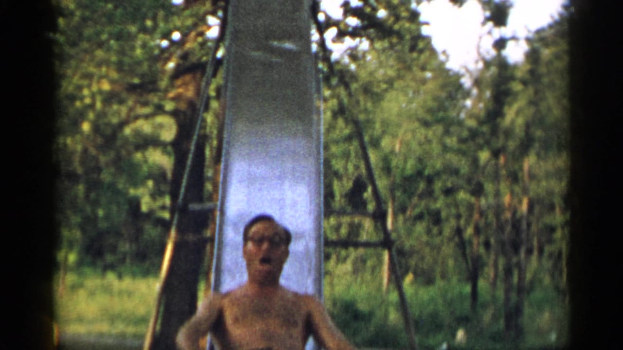 The man nears the bottom of the tall metal slide, with glasses and facial features visible in the 1954 New York home-movie frame.