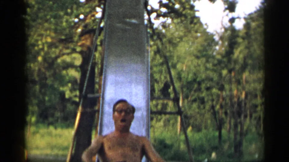 The man nears the bottom of the tall metal slide, with glasses and facial features visible in the 1954 New York home-movie frame.