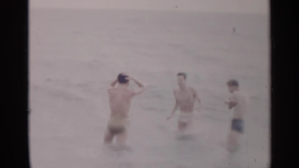 In 1954 New York beach footage, three young men in swim trunks stand in soft-focus ocean surf as one adjusts a cap.