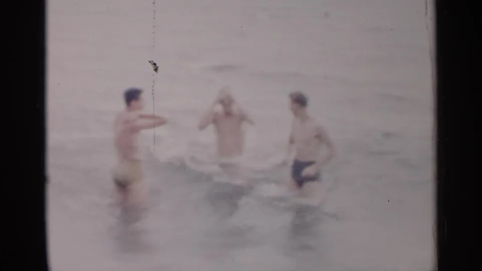 Three young men in swim trunks play in shallow New York ocean surf, partly obscured by faded color and film wear.