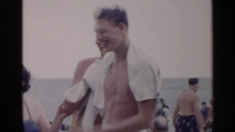 A smiling young man with a white towel over his shoulder stands near other beachgoers on a New York shoreline.
