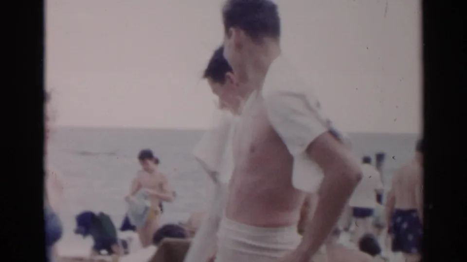 Two young men with towels stand on a crowded New York beach, with the ocean and seated beachgoers behind them.