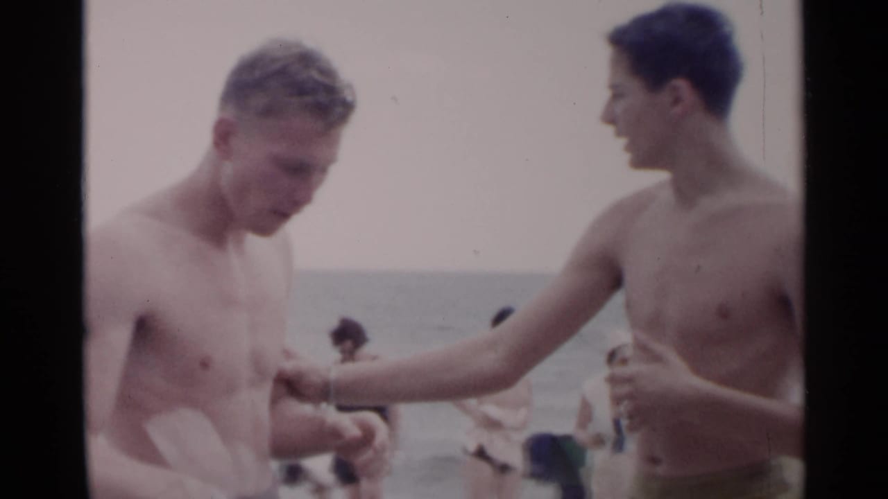 Two shirtless young men stand on a beach near the waterline.