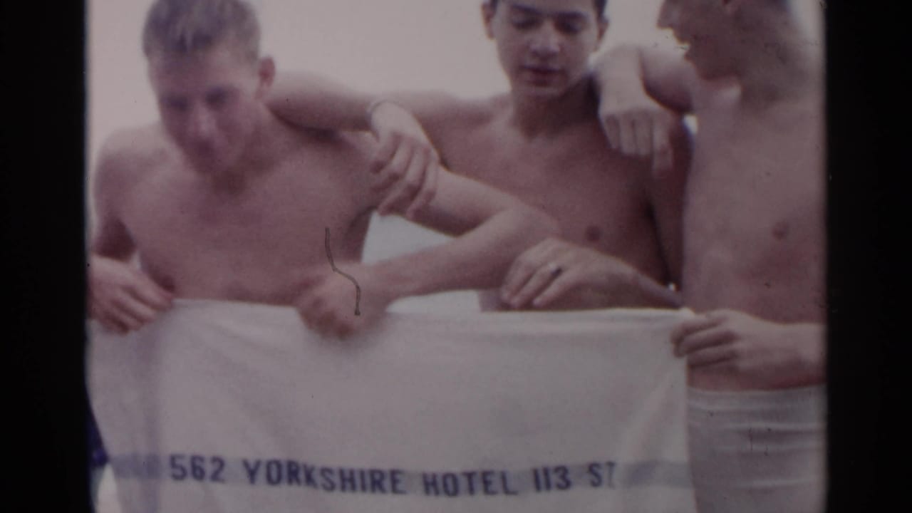 Three young men hold up a white towel with hotel text.